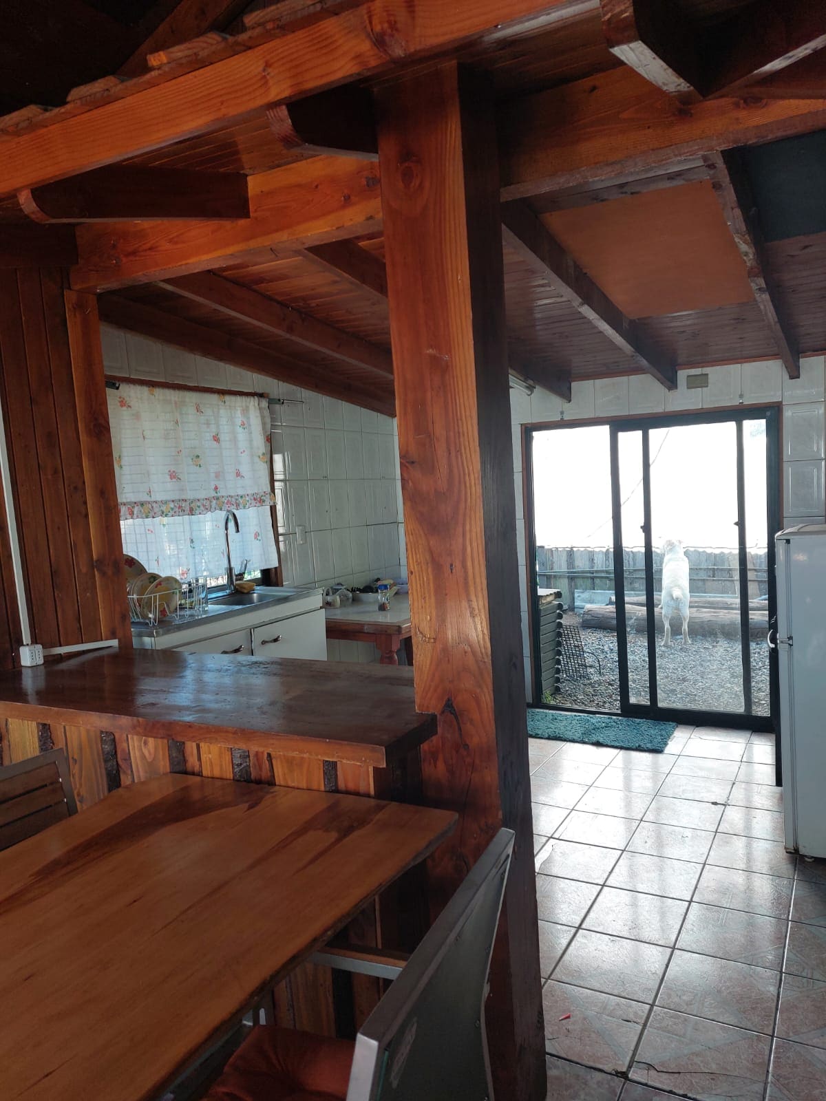 An open kitchen area showcases wooden beams and warm tones, with a dining table positioned in the foreground. A glass door leads to an outdoor space, allowing natural light to enter, while a view of a patio area is visible through the door.