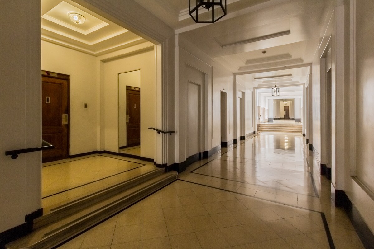A well-lit hallway features smooth tile flooring and high ceilings with ornate lighting fixtures. Doorways are visible on both sides, leading to various rooms. The design is modern and elegant, with a neutral color palette enhancing the sense of spaciousness.