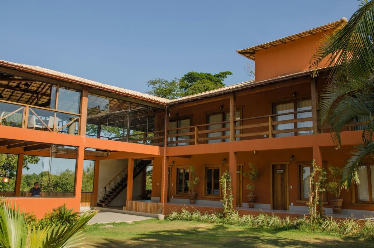 The charming farmhouse features a two-story structure with a warm orange exterior. A spacious terrace is visible, complemented by large glass windows. Lush greenery surrounds the building, enhancing the inviting outdoor space that includes neatly arranged pathways and plants.