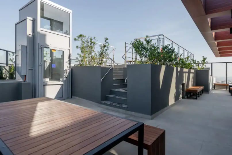 A rooftop area is showcased, featuring several planters with greenery and a staircase leading to an upper level. Wooden tables are positioned on a gray surface, providing space for outdoor gatherings and relaxation. The surrounding area is bathed in natural light.