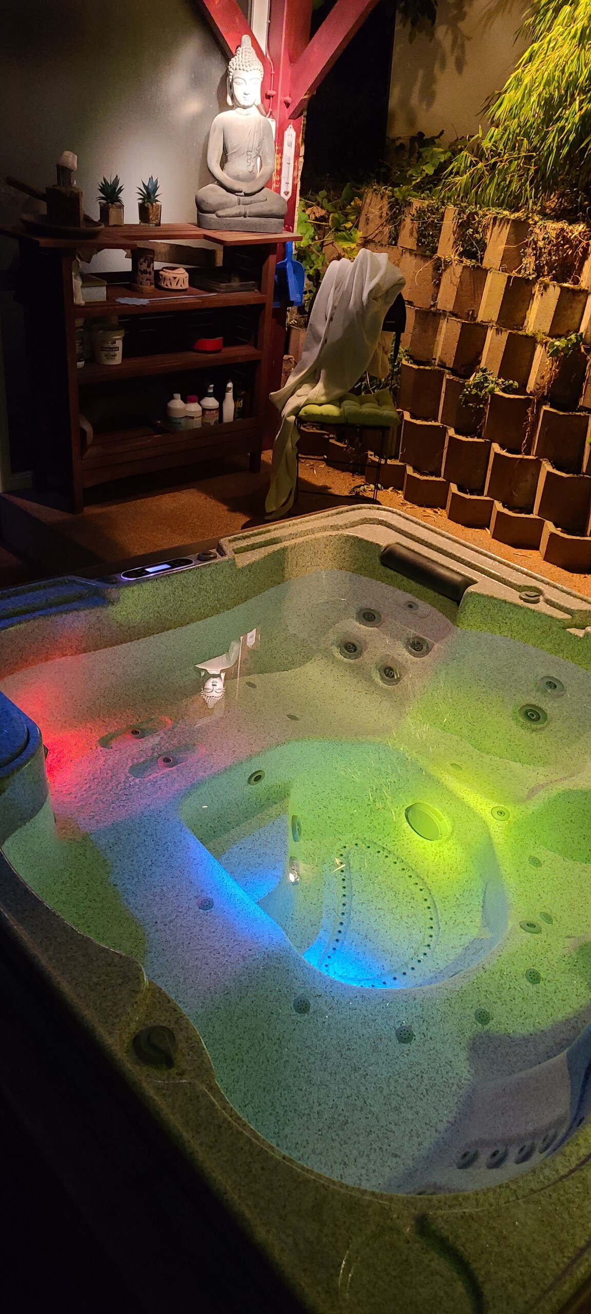 A relaxing hot tub is showcased, with integrated LED lights casting vibrant colors in a calming atmosphere. A wooden shelf within view holds various toiletries, while a serene Buddha statue adds a tranquil touch to the surrounding area.