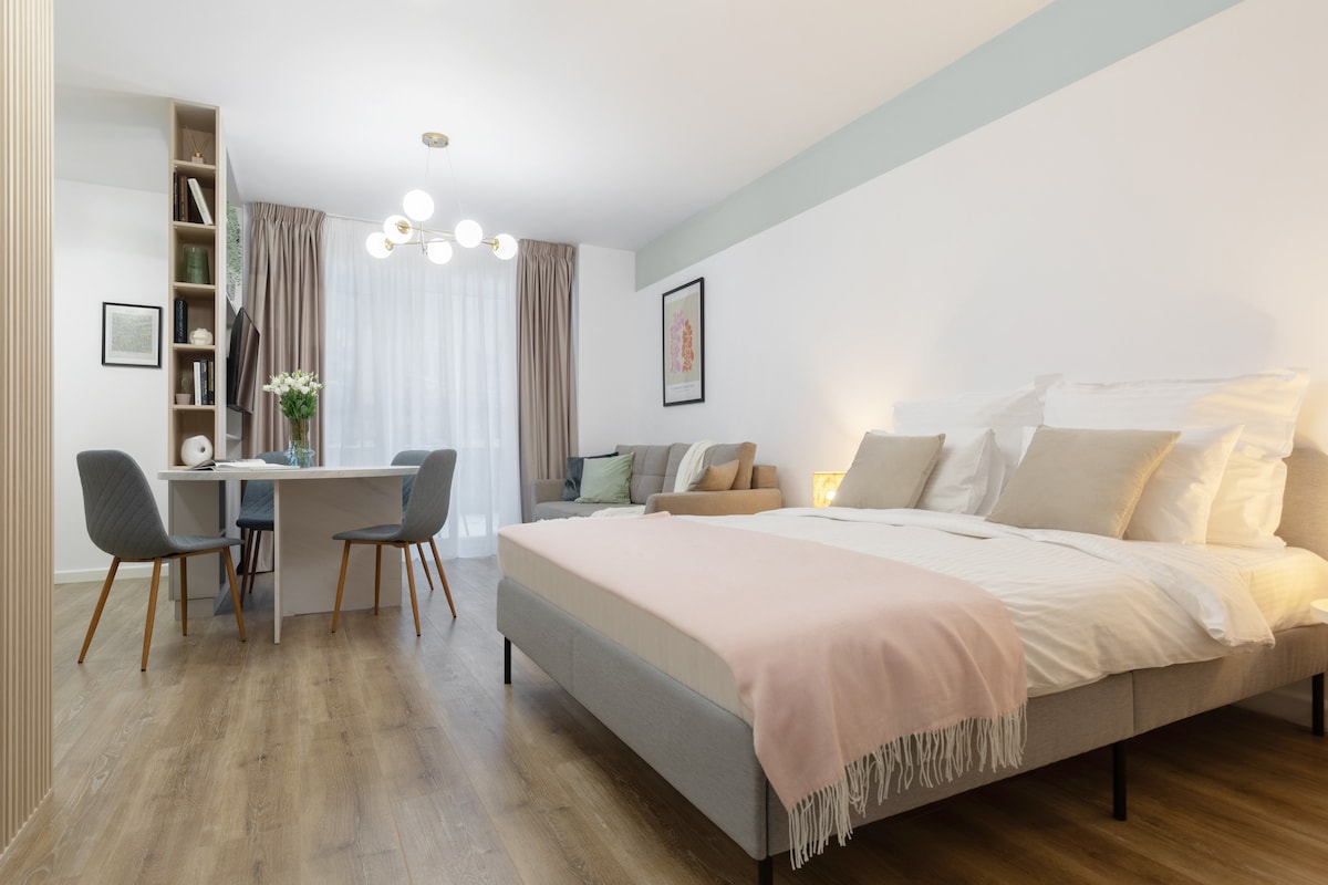 A spacious sleeping area features a large, neatly made bed with soft linens and accent pillows. A dining table with chairs is positioned nearby, while a cozy seating area is visible, complemented by soft lighting and large windows allowing natural light to fill the room.