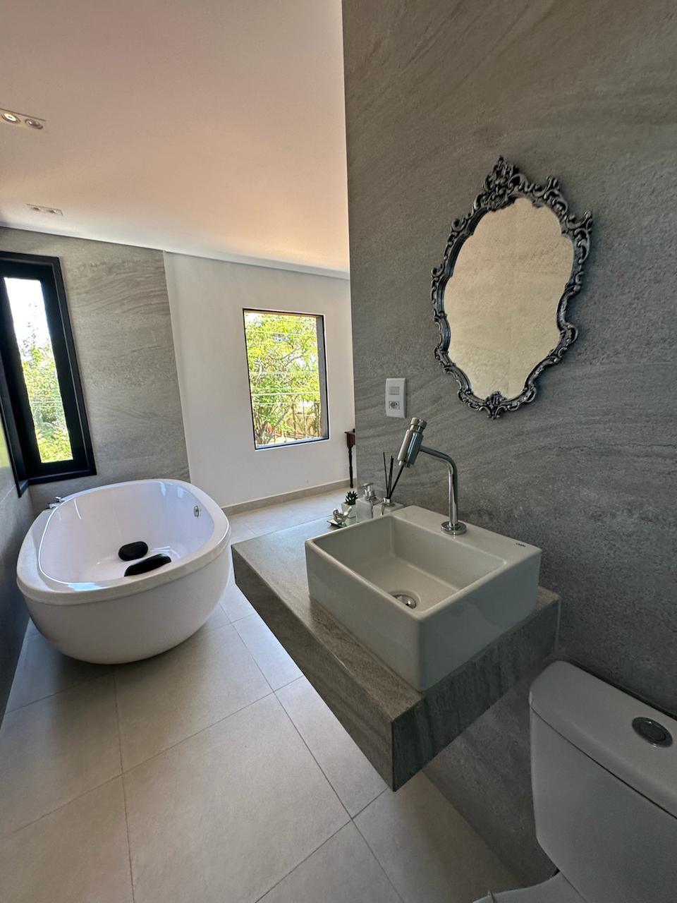 A modern bathroom features a freestanding bathtub alongside a sleek sink with a minimalistic faucet. Natural light enters through a large window, complementing the gray stone walls. A decorative mirror is mounted on the wall, adding an elegant touch to the space.