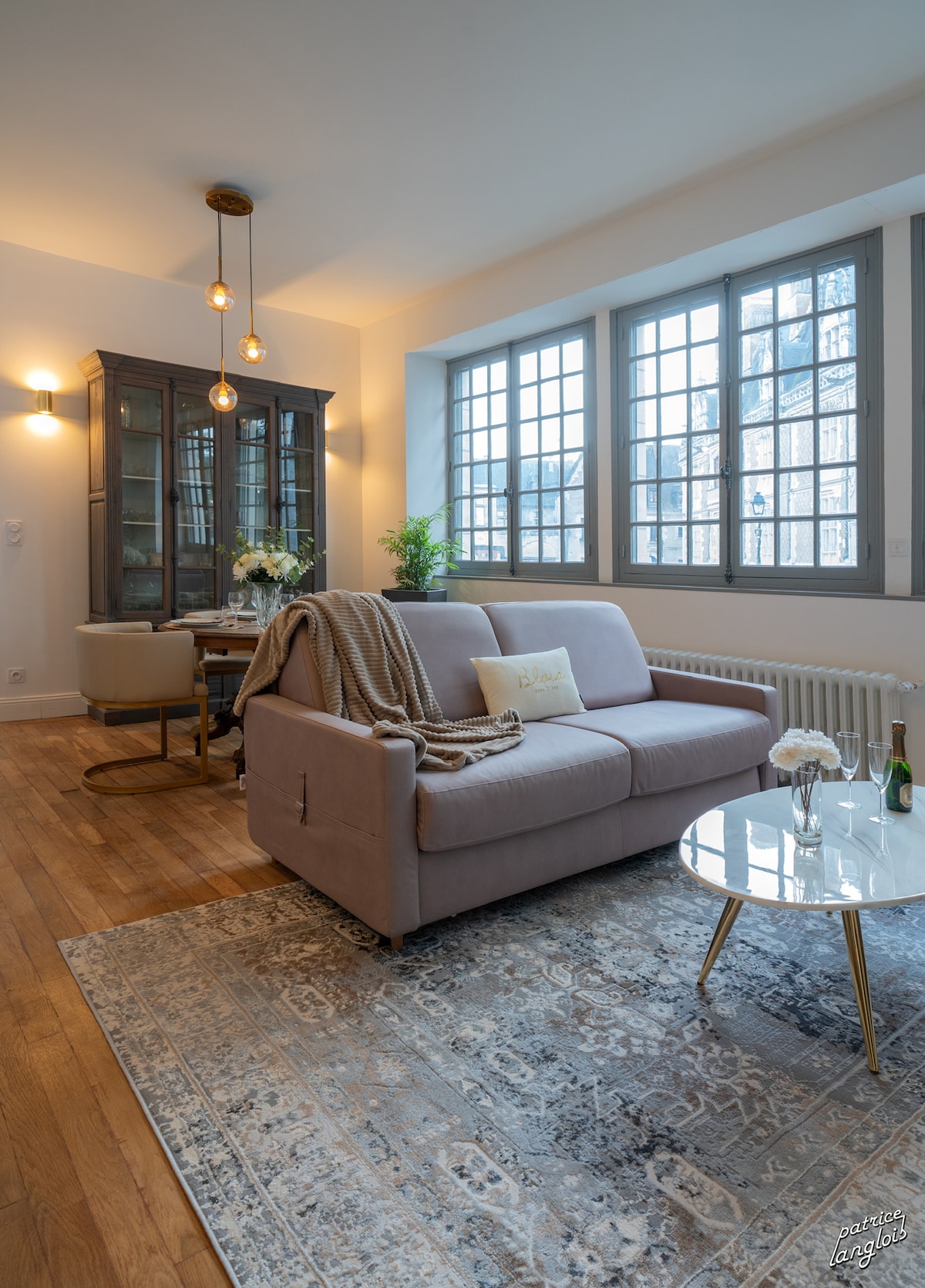 A light-filled living area showcases a comfortable sofa draped with a soft throw. A round table with dining chairs is positioned nearby, while large windows allow natural light to illuminate the space. A decorative rug adds warmth to the wooden flooring.