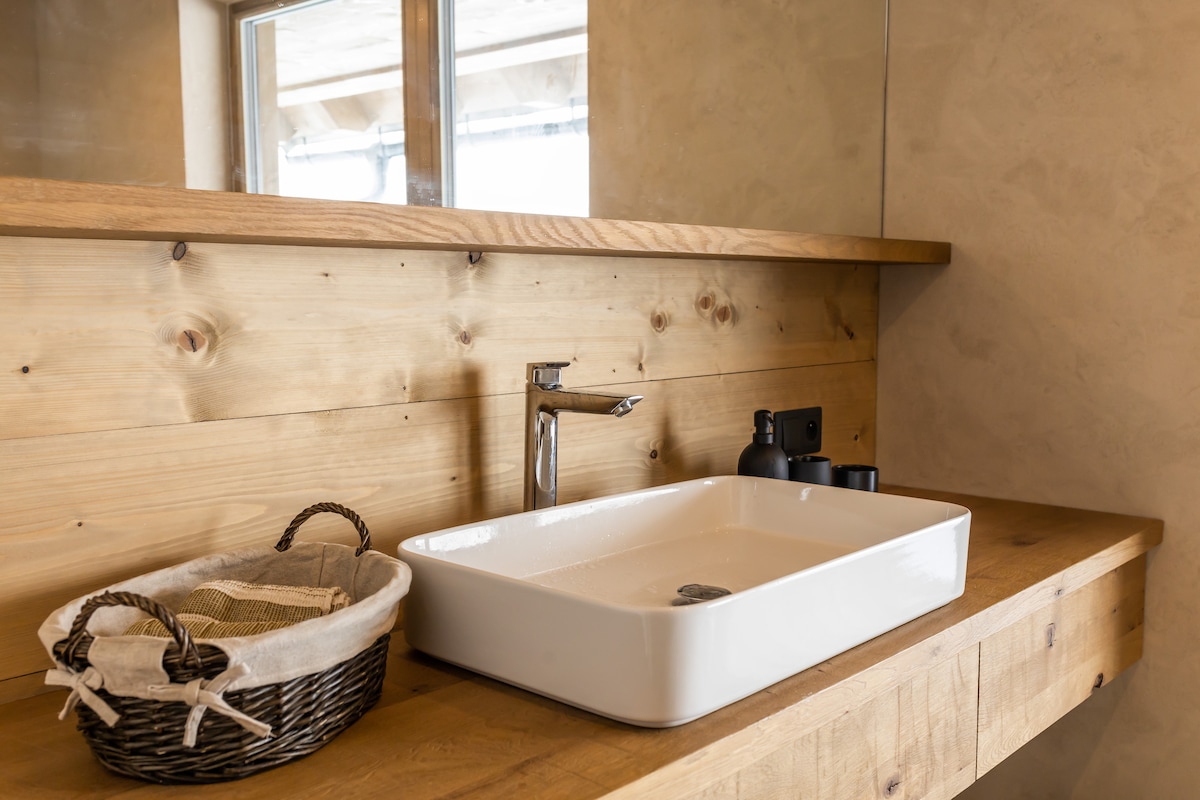 A wooden countertop features a sleek white sink, complemented by a modern faucet. A woven basket rests beside the sink, filled with essential toiletries, while a mirror reflects natural light, enhancing the space's clean and functional design.