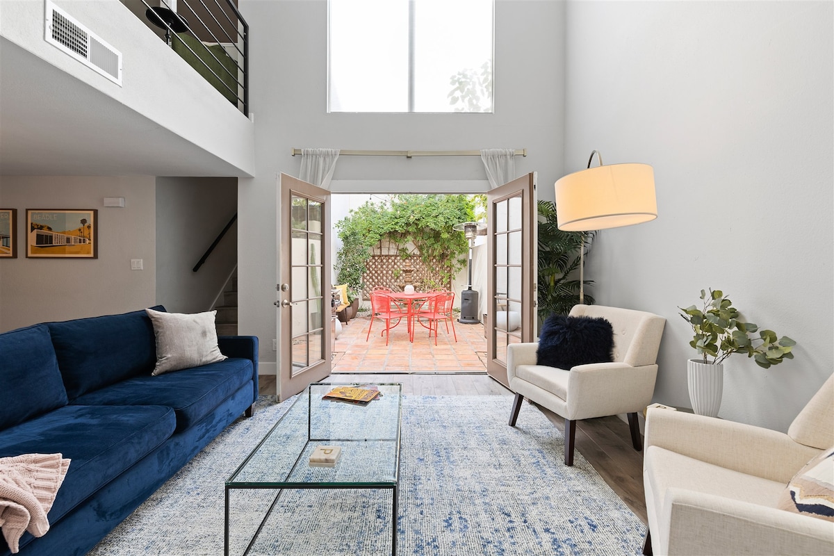 A bright living area features a blue sofa and two light-colored chairs surrounding a glass coffee table. French doors open to a patio, bringing natural light and outdoor views into the space. A rug adds warmth, while greenery enhances the ambiance.