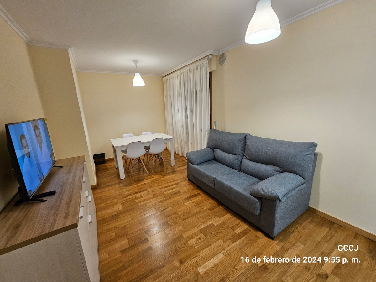 A living space features a comfortable sofa and a light wooden dining table surrounded by four chairs. A television is placed on a cabinet, and large curtains cover a window, allowing ambient light to fill the room. Warm wooden flooring complements the neutral wall tones.