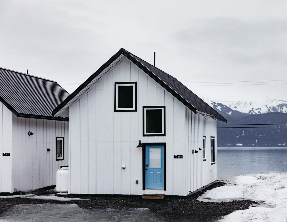 A modern seaside cottage is presented with a bright blue front door. The exterior features white paneling and large windows, allowing for ample natural light. Snow can be seen on the ground, and the view of a calm body of water and distant mountains is visible in the background.