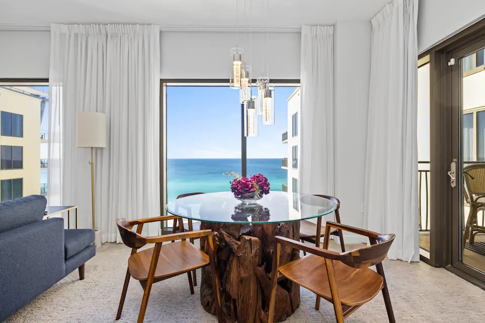 A dining area features a round glass table supported by a wooden base, surrounded by four chairs. Large windows provide a view of the beach and ocean, while soft curtains frame the light-filled space, enhancing the serene atmosphere.