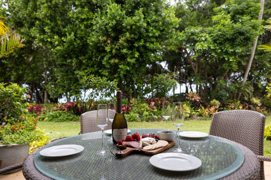 A dining table is set outdoors, featuring white plates, champagne glasses, and a platter of strawberries and cheese. Lush greenery and colorful foliage create a serene backdrop, enhancing the outdoor dining experience.
