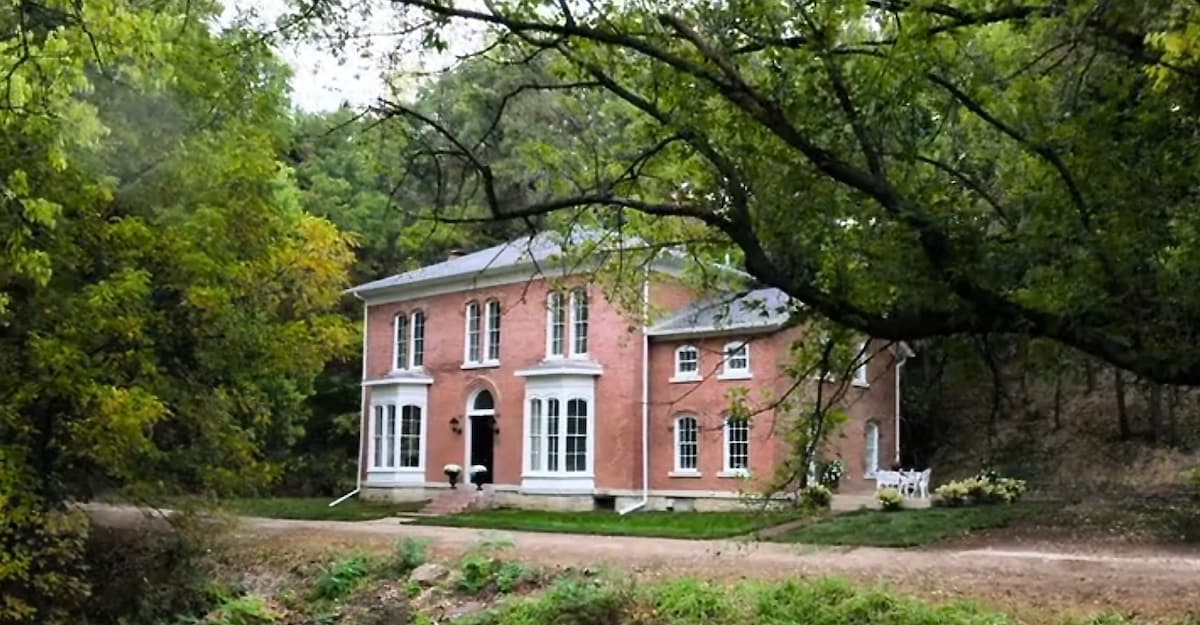 The historic Horn Hollow estate is depicted, showcasing its elegant brick facade framed by lush trees. Large windows enhance the structure's classic architecture, inviting ample natural light. A well-maintained lawn surrounds the home, providing a serene country setting.