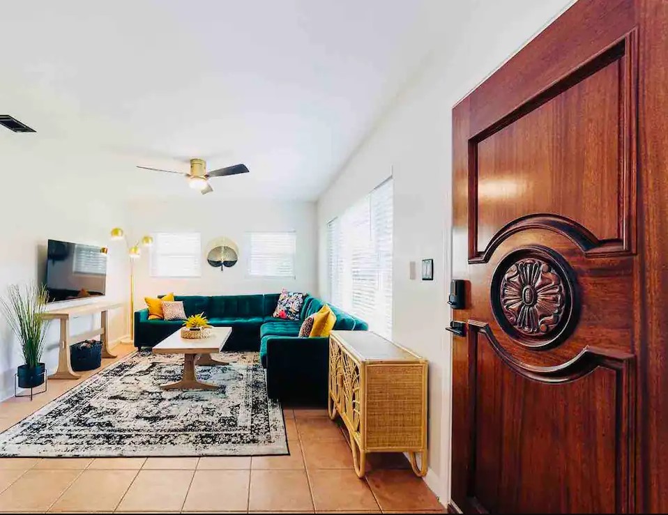 A welcoming living area features a dark green sectional sofa adorned with colorful cushions. A decorative area rug lies on the tiled floor. Natural light filters through multiple windows, and a wooden door with an intricate design stands prominently in the foreground.
