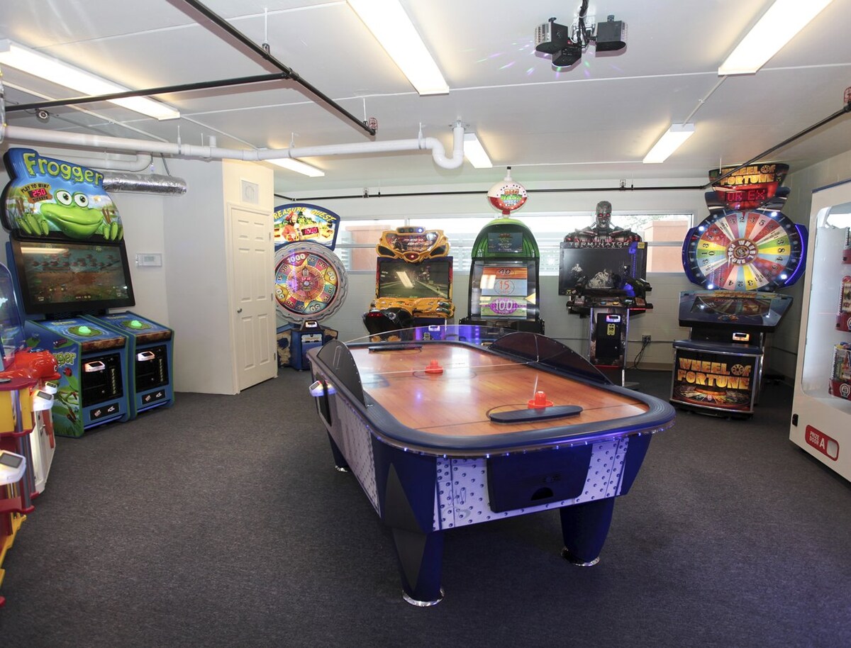 A game room features a variety of arcade machines, including classic titles and modern games. An air hockey table occupies the center, surrounded by vibrant machines displaying colorful graphics and illuminated screens. The space is carpeted, creating a comfortable environment for entertainment.