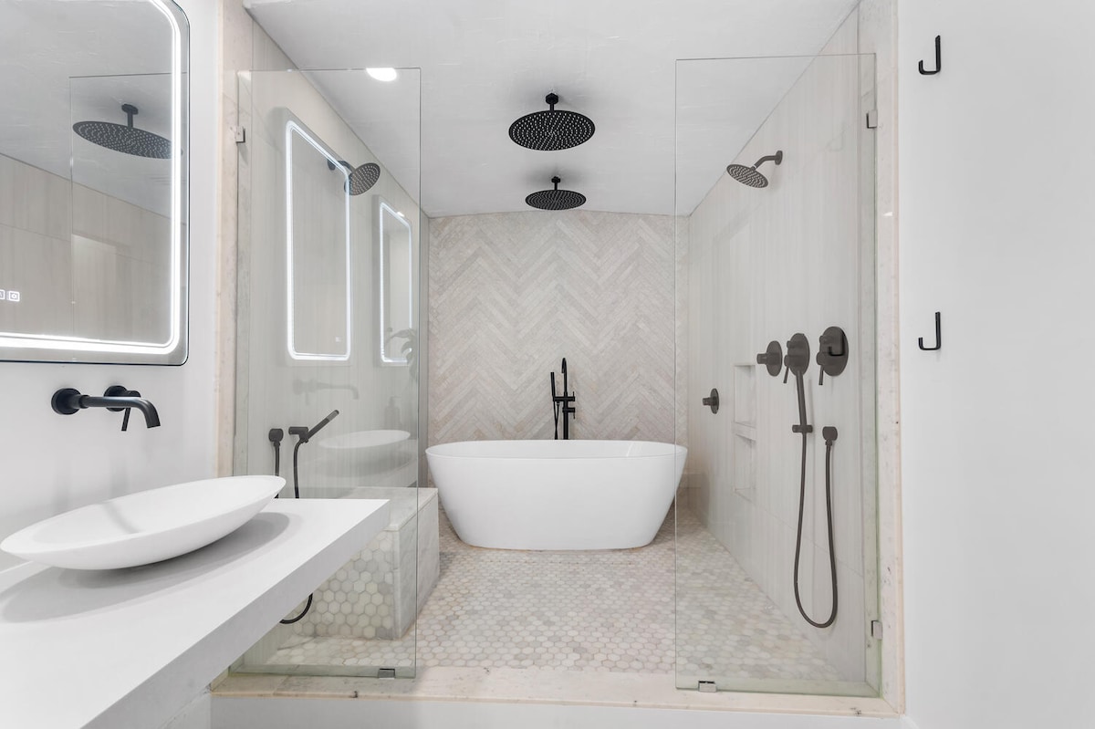 A modern bathroom features a freestanding tub surrounded by glass panels, enhancing a sleek aesthetic. Dual rainfall showerheads complemented by minimalist fixtures are visible. Soft lighting from backlit mirrors enhances the serene environment, while a stylish basin is positioned on a floating vanity.