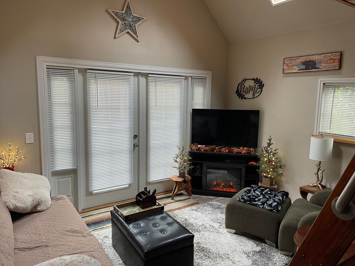 A cozy living area features a comfortable couch and armchair, arranged around an electric fireplace beneath a flat-screen TV. Natural light filters through the windows adorned with blinds. Decorative elements include small trees and a star, adding a touch of charm to the space.
