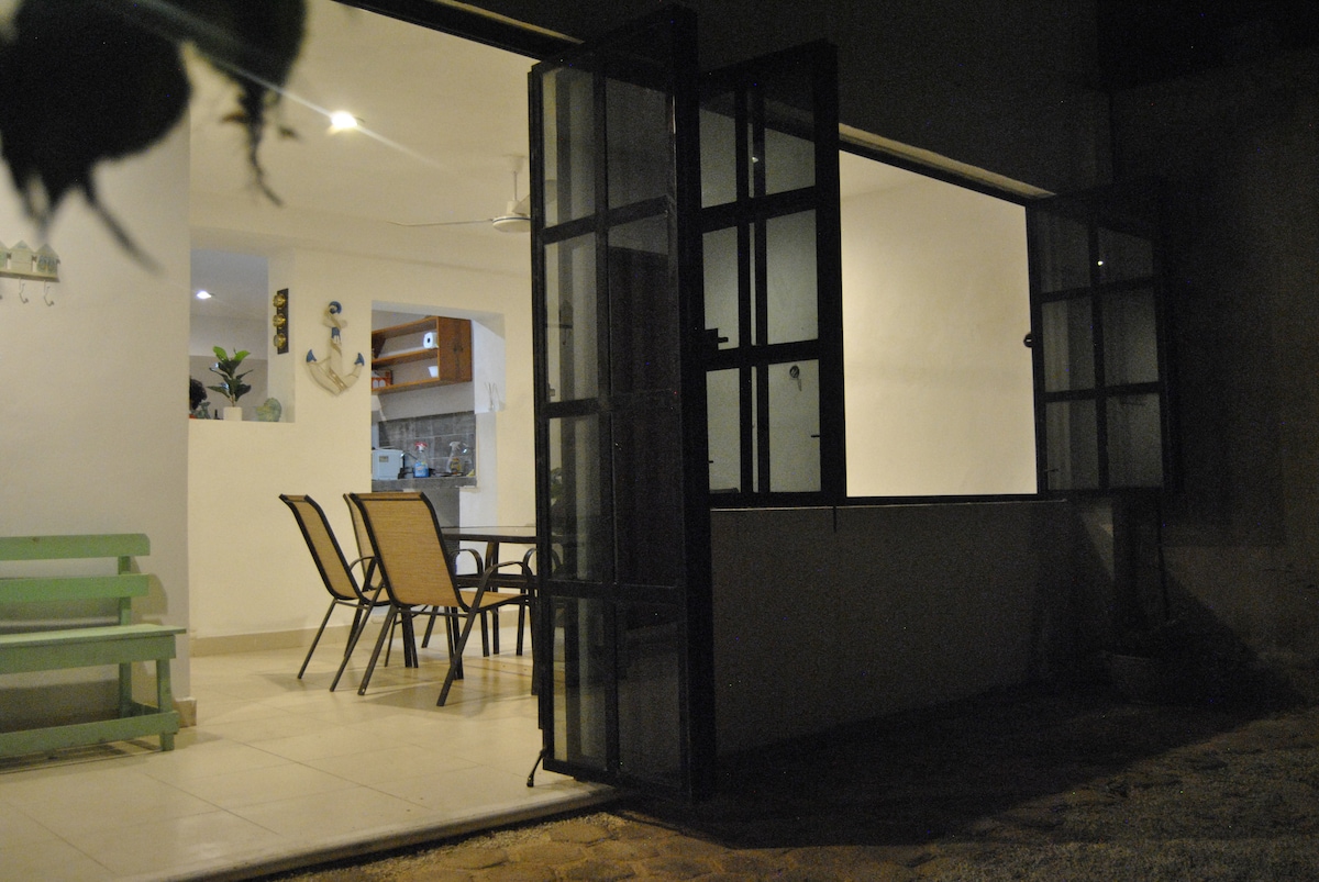 Large glass doors are open, connecting an indoor dining area with outdoor space. A simple wooden table is surrounded by several chairs. The adjacent kitchen area is partially visible, illuminated softly, while the walls reflect a neutral color palette.