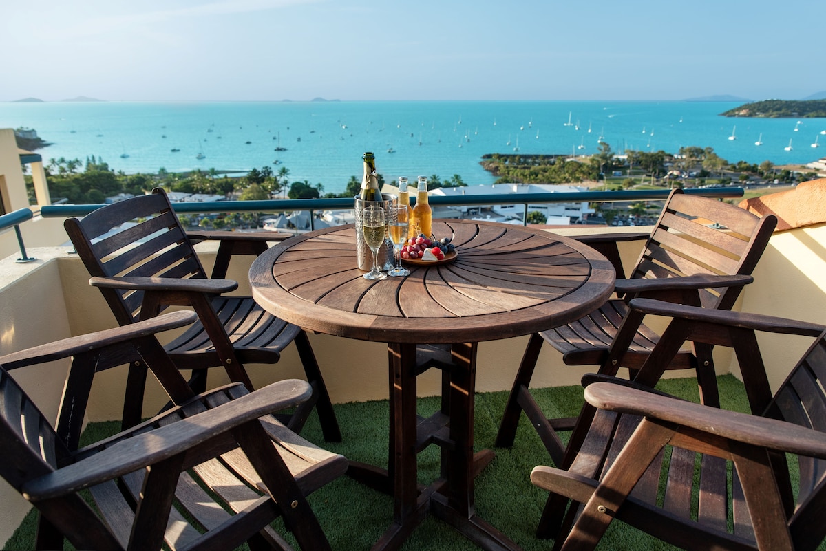A large round wooden table is surrounded by four sturdy chairs on a private balcony. Glasses filled with beverages and a platter of fresh fruit are arranged on the table, with the expansive ocean and distant sailboats visible in the background.