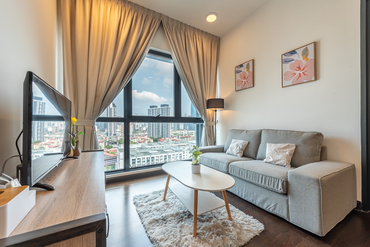A contemporary living area features a gray sofa paired with a low coffee table, positioned by a large window that offers views of the city skyline. Soft curtains frame the window, and a TV is mounted on a media console, creating a relaxed atmosphere.