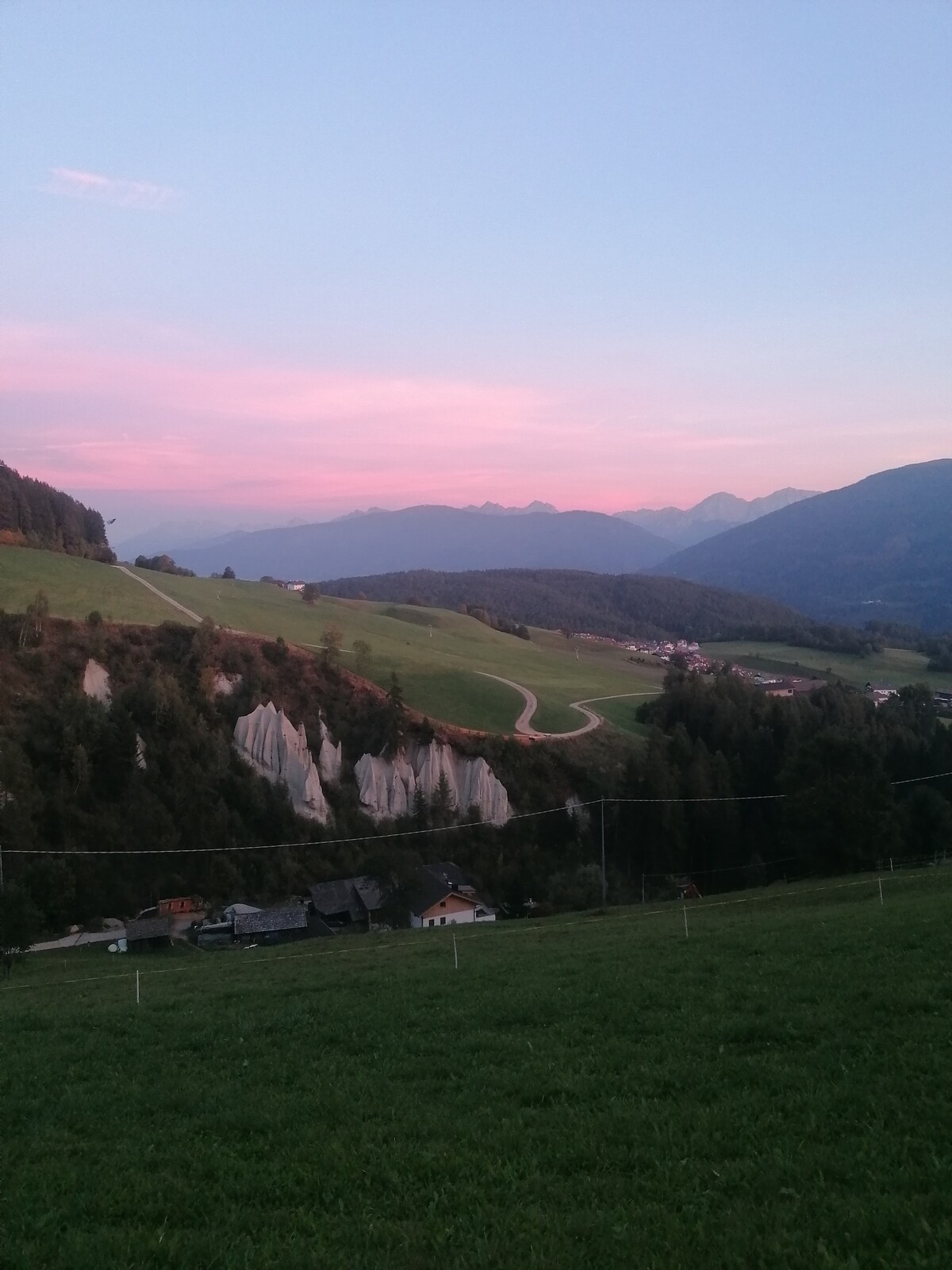 A serene landscape at dusk showcases gentle hills and distant mountain peaks under a soft pink sky. The foreground features lush green grass, while white earth pyramids are visible along the hillside. Small buildings are scattered in the valley below, adding to the tranquil scene.