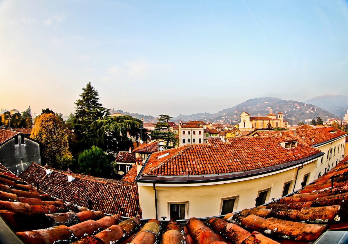 The panoramic view showcases terracotta rooftops under a soft sky, with green trees and historic buildings in the background. The distant hills of Brescia create a scenic and tranquil landscape, inviting exploration of the area’s rich heritage.