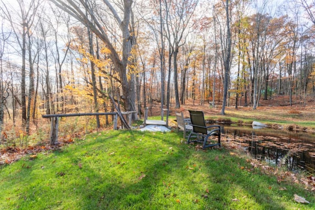 A serene outdoor area is shown alongside a pond, featuring a grassy patch bordered by trees with autumn foliage. Two metal chairs are positioned near a wooden railing, providing a peaceful spot to enjoy nature.
