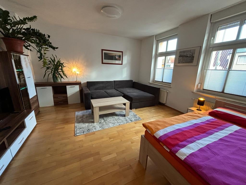A comfortable living area is presented, featuring a dark sectional sofa and a light coffee table atop a soft rug. Natural light enters through large windows, highlighting a wooden cabinet and a plant positioned nearby. A colorful bed is visible on the right side of the room.