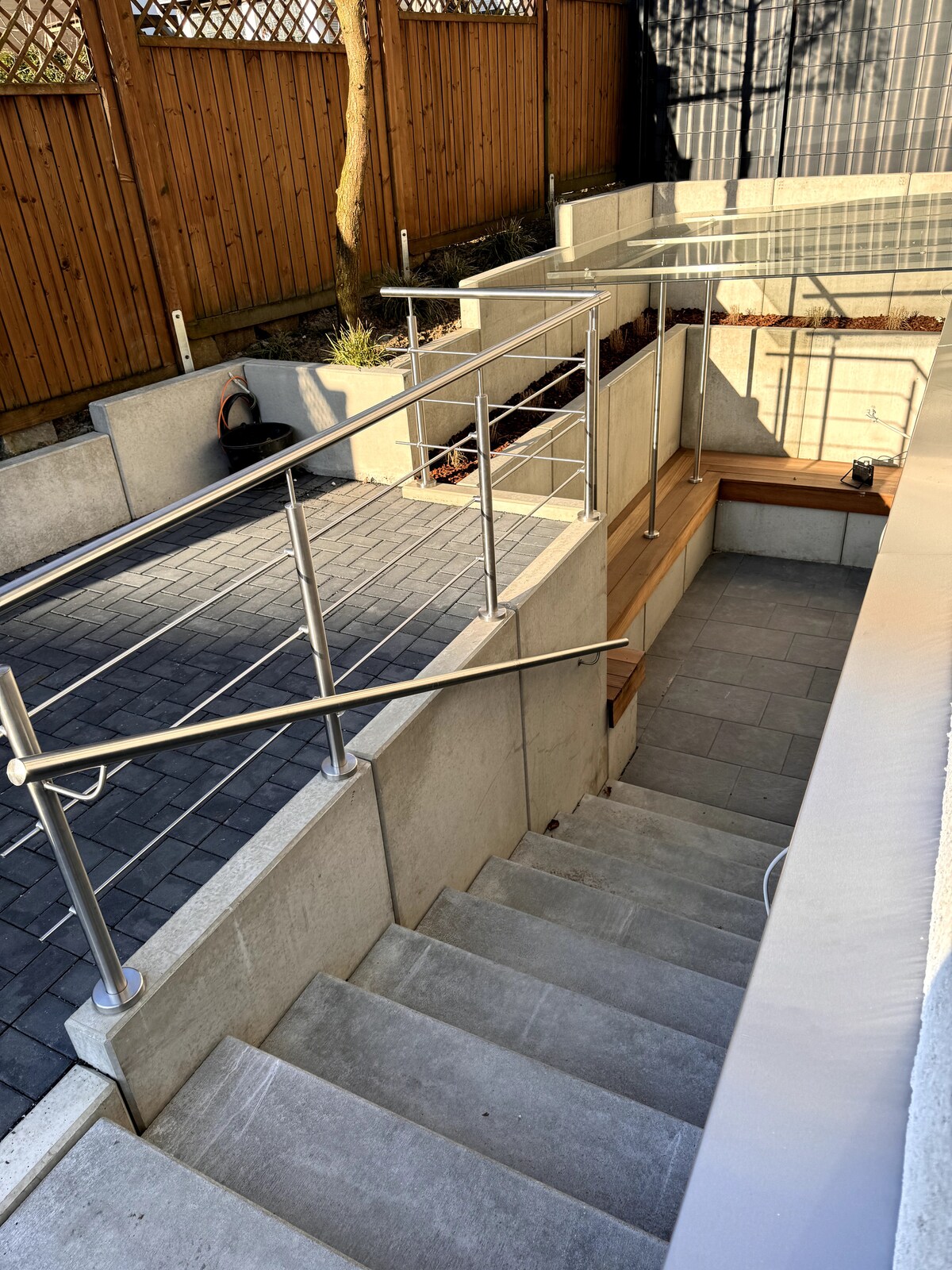 A set of smooth concrete stairs leads down into a sunlit outdoor area. Railing made of sleek metal provides safety, while paved pathways and wooden seating areas create a structured yet inviting space. Wooden fencing borders the area, adding privacy.