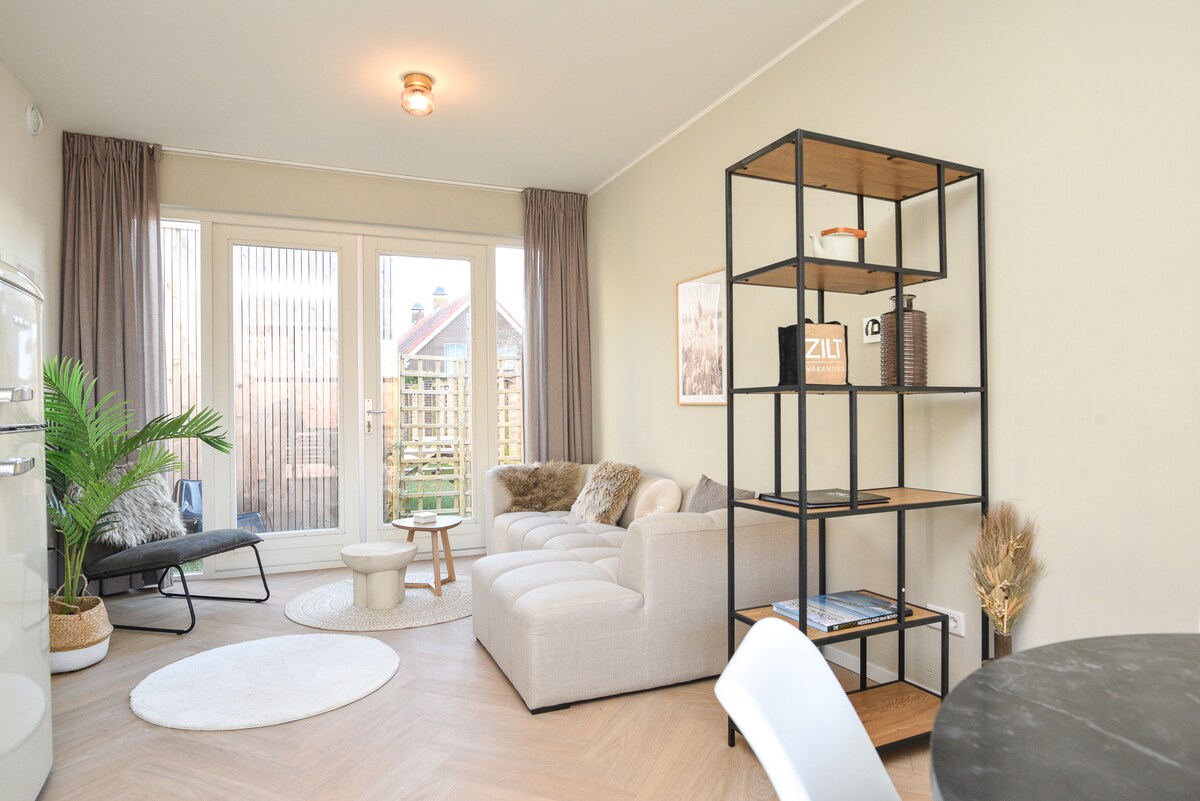 The living area features a light-toned sofa, a round coffee table, and a minimalist bookshelf. Large glass doors provide access to the outdoor space, inviting natural light into the room. A cozy area rug and a plant add to the comfortable atmosphere.