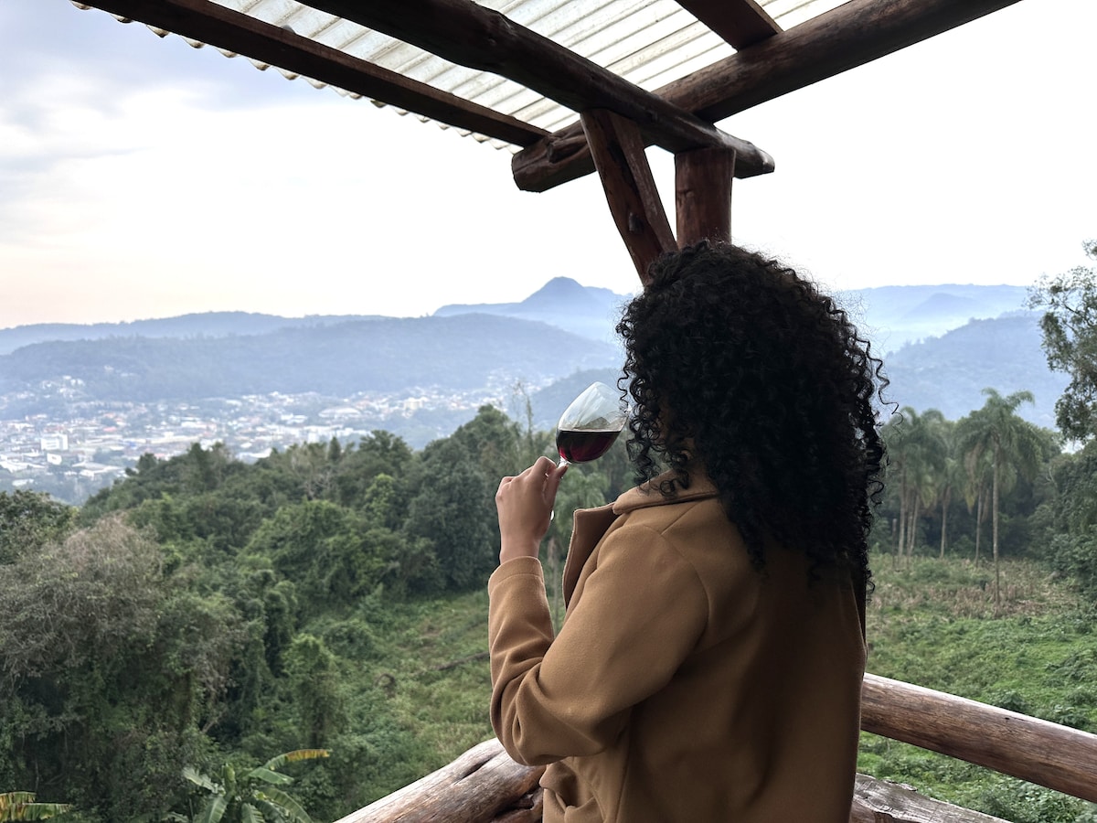 A woman is seen holding a glass of red wine while gazing at a view of the city and surrounding mountains. The natural landscape features lush greenery and distant hills, with a wooden structure providing a cozy outdoor seating area.
