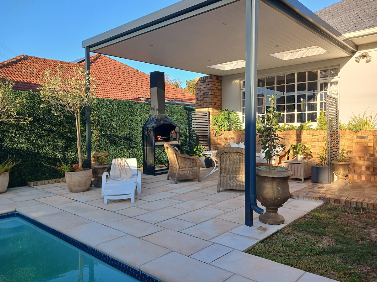 An outdoor patio area is set beside a swimming pool, featuring a large barbecue fireplace and comfortable seating arrangements. Potted plants and greenery surround the space, while a shaded roof provides shelter. Natural stone tiles lead to the pool, creating an inviting relaxation spot.