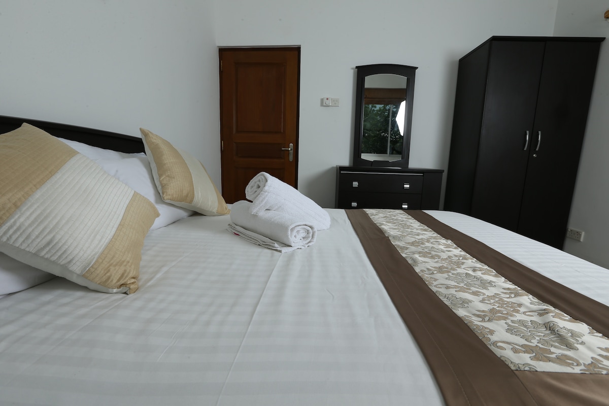 A spacious double bed is adorned with decorative pillows and a patterned bed runner. Neatly rolled towels are placed on the sheets. A mirror and dresser occupy one side of the room, complemented by a dark-colored wardrobe against the wall.