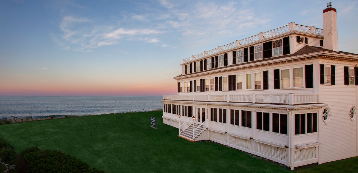 The historic hotel building features a white facade and multiple windows, with a balcony overlooking the expansive green lawn and ocean. The soft colors of a sunset are visible in the background, adding warmth to the serene coastal scene.