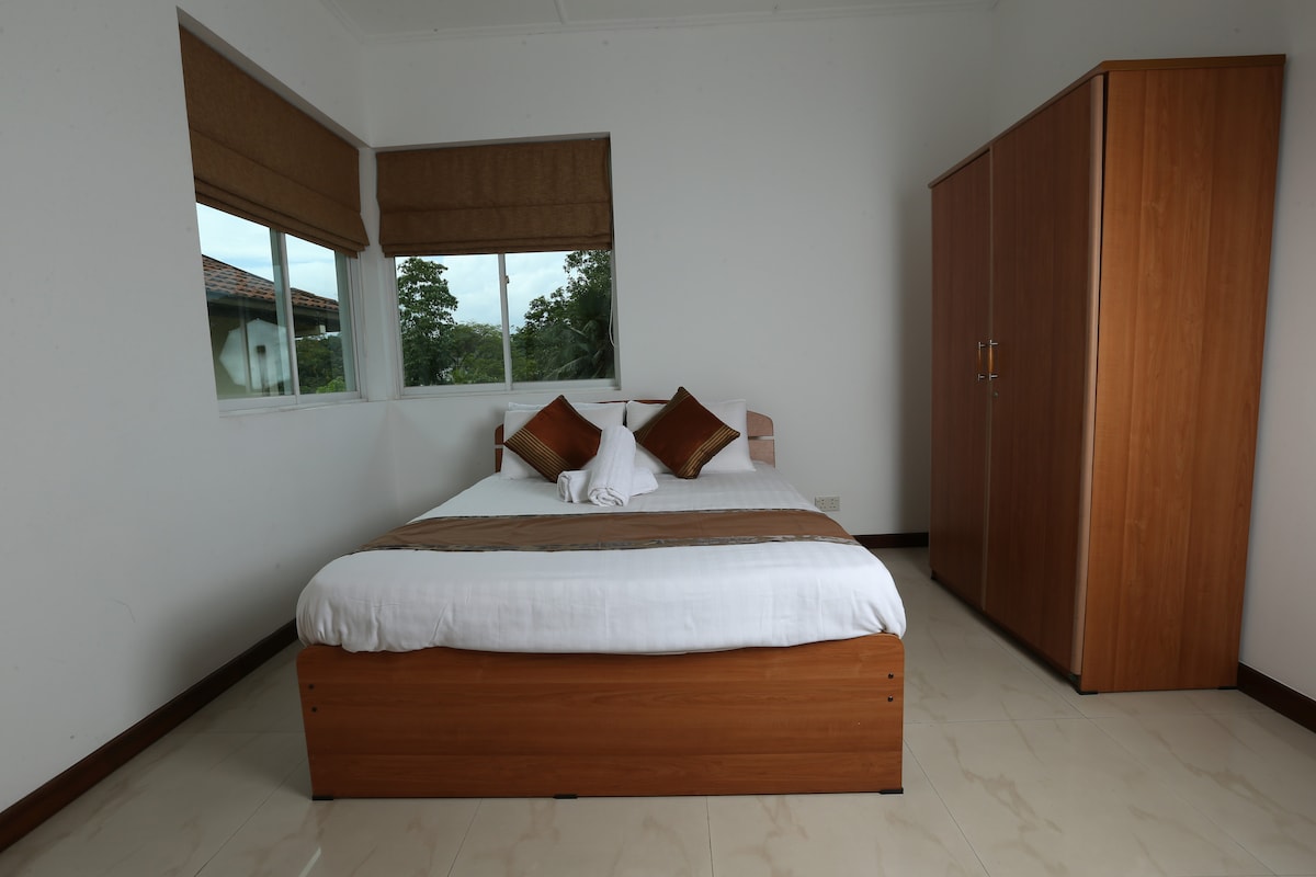 A spacious bedroom is featured with a large bed dressed in white linens, complemented by decorative pillows. Natural light enters through two windows adorned with neutral-toned shades. A wooden wardrobe stands nearby, providing ample storage space.