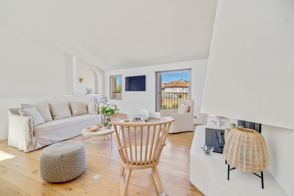 A bright living area features a comfortable sofa adorned with neutral cushions and a decorative pouf. Large windows allow natural light to fill the space, and a television is mounted on the wall. A coffee table sits in the center, complemented by a small floral arrangement.