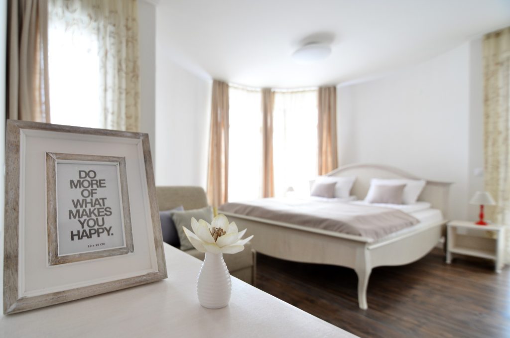 A bright and spacious room features a large bed with soft linens and two pillows. Natural light fills the space through multiple windows adorned with light curtains. A small desk displays a minimalistic framed quote and a vase with a white flower, enhancing the serene ambiance.
