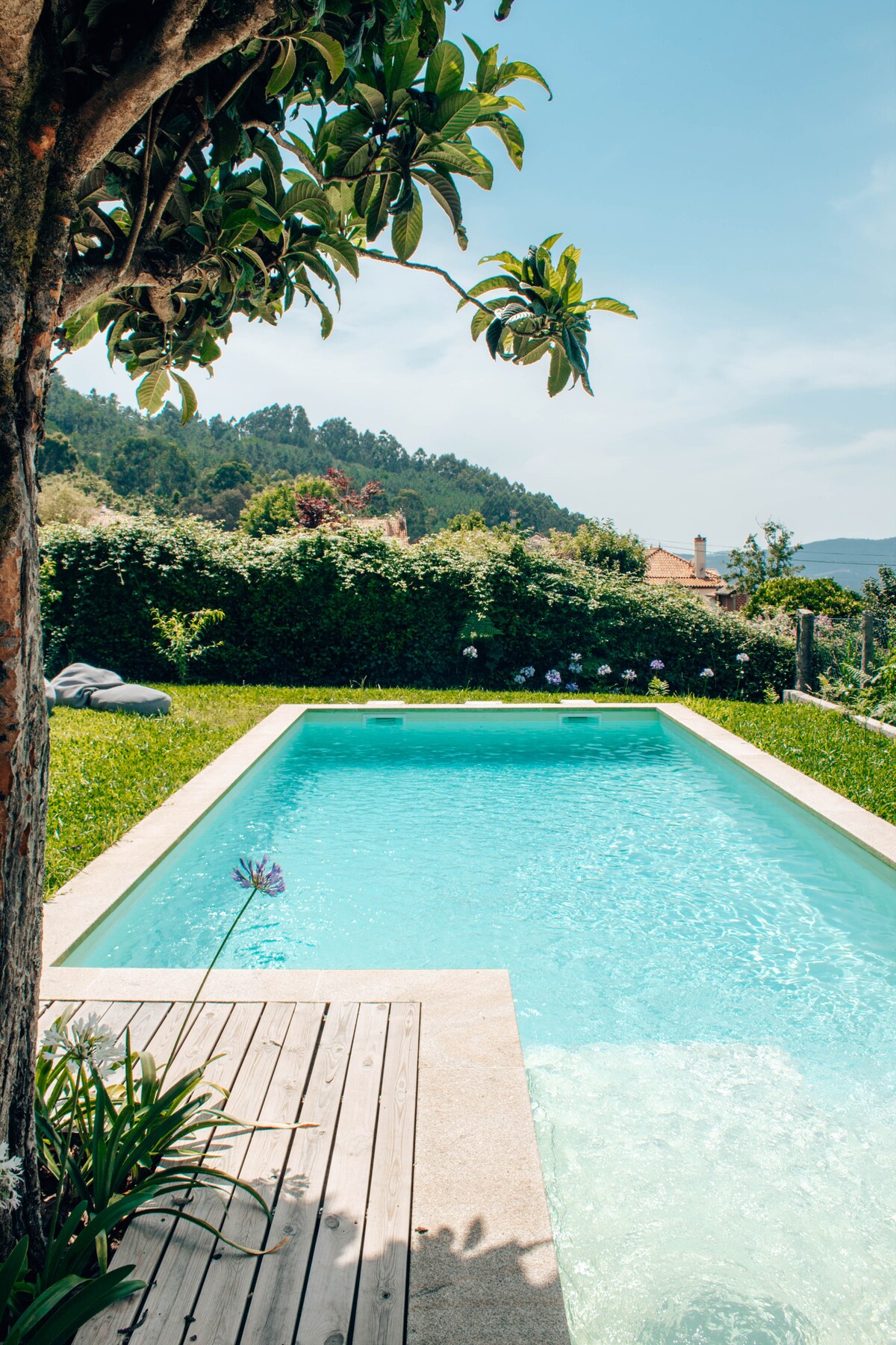 A serene swimming pool is surrounded by lush greenery, featuring a well-maintained deck on one side. The clear water reflects sunlight, while a single flower adds a touch of color to the garden landscape. The backdrop includes gently rolling hills, providing a peaceful view.