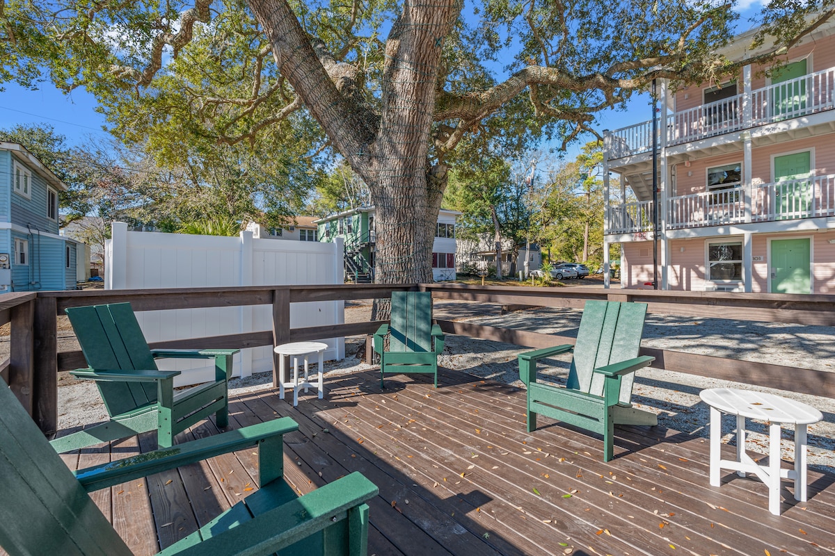A shaded outdoor deck features green Adirondack chairs arranged around a small white table, surrounded by gravel and a large tree. The area provides a tranquil space for relaxation, with views of the adjacent building and parking area.