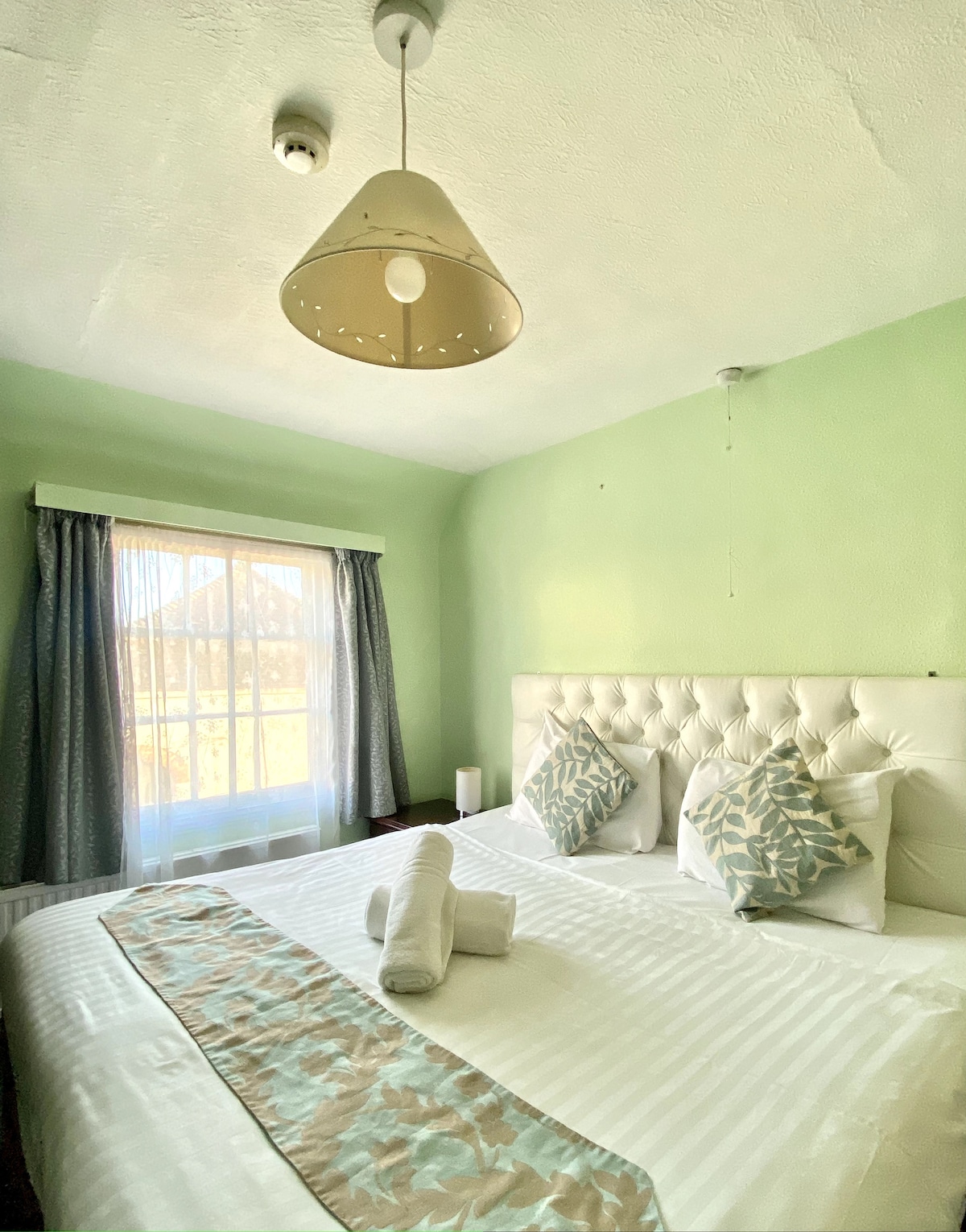 A well-lit bedroom features a spacious bed adorned with decorative pillows and a lightly patterned throw. Natural light enters through a large window dressed with sheer curtains, while a modern pendant light provides additional illumination.