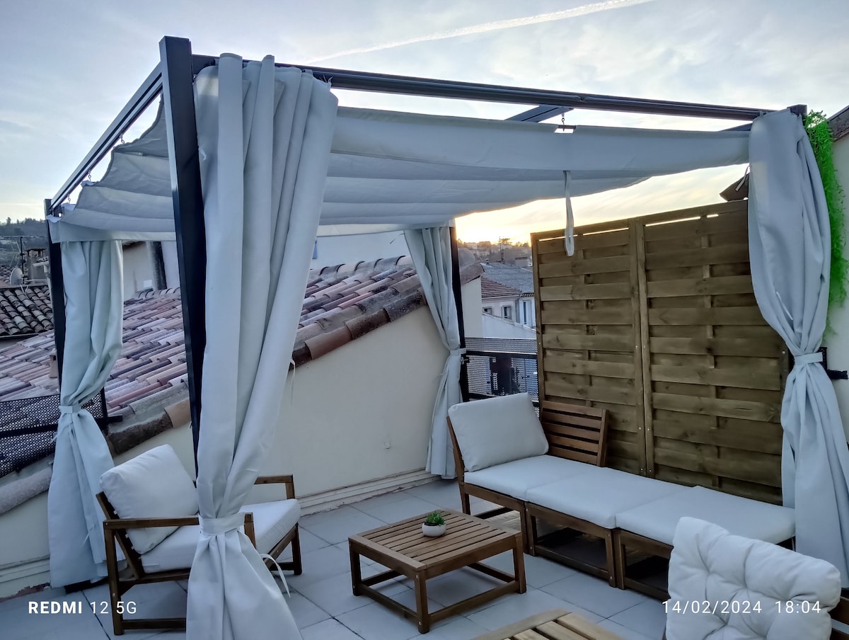 A serene terrace features wooden furniture and light-colored cushions. The space is partially sheltered with white drapes, providing a peaceful spot for relaxation. A small potted plant is centered on a table, and a view of rooftops is visible beyond, illuminated by soft evening light.