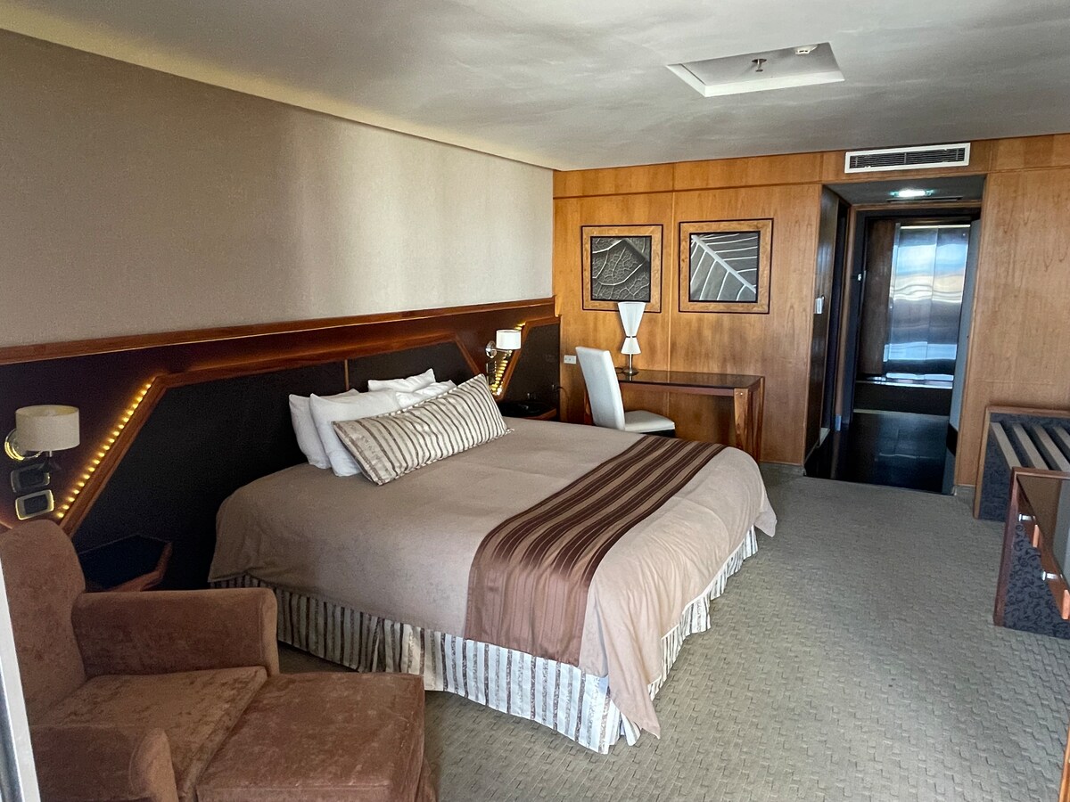 A spacious room features a large bed dressed in neutral-toned linens. Wood paneling and soft lighting create a cozy feel. A desk and chair are positioned next to the bed, while a plush armchair offers additional seating. Visible doors lead to an adjacent area.