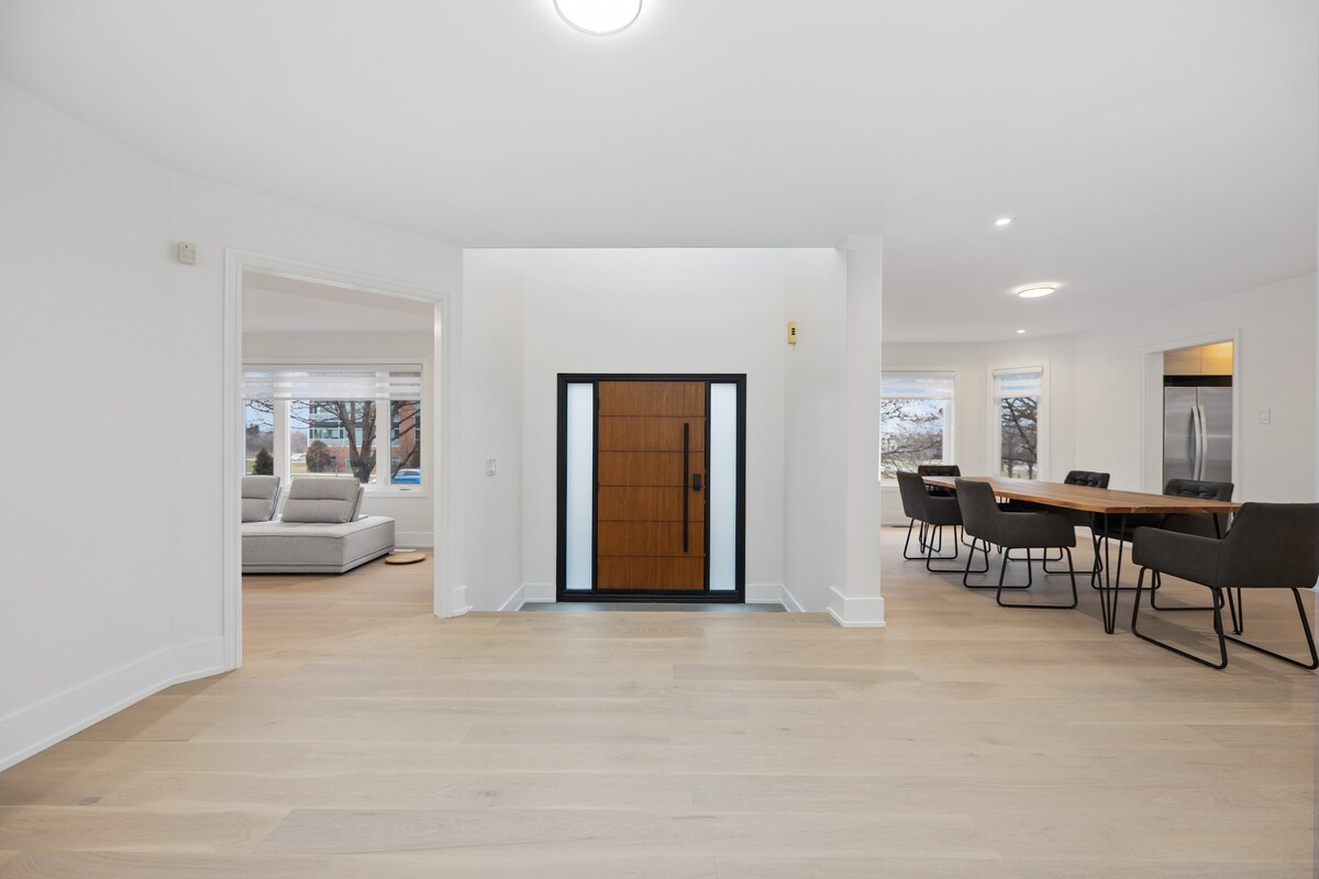 A bright entryway is highlighted by a modern front door featuring a large glass panel. The smooth wooden floor extends to an open dining area, furnished with a long table and stylish chairs. Natural light fills the space from large windows in the adjacent living room.