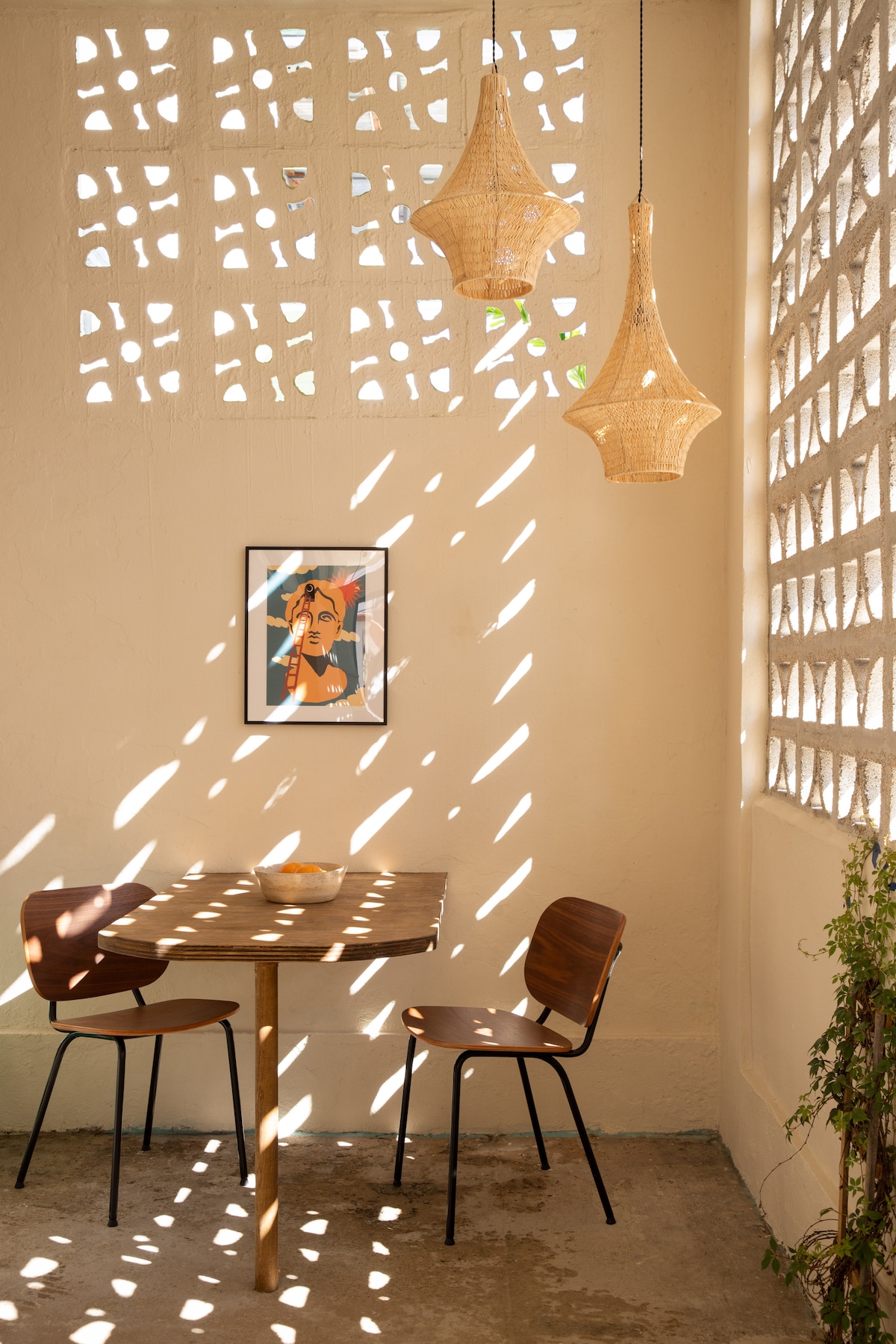A dining area features a wooden table accompanied by two chairs, illuminated by soft light streaming through uniquely patterned wall panels. Woven pendant lights hang above, highlighting a framed artwork on the adjacent wall, while a potted plant adds a touch of greenery.