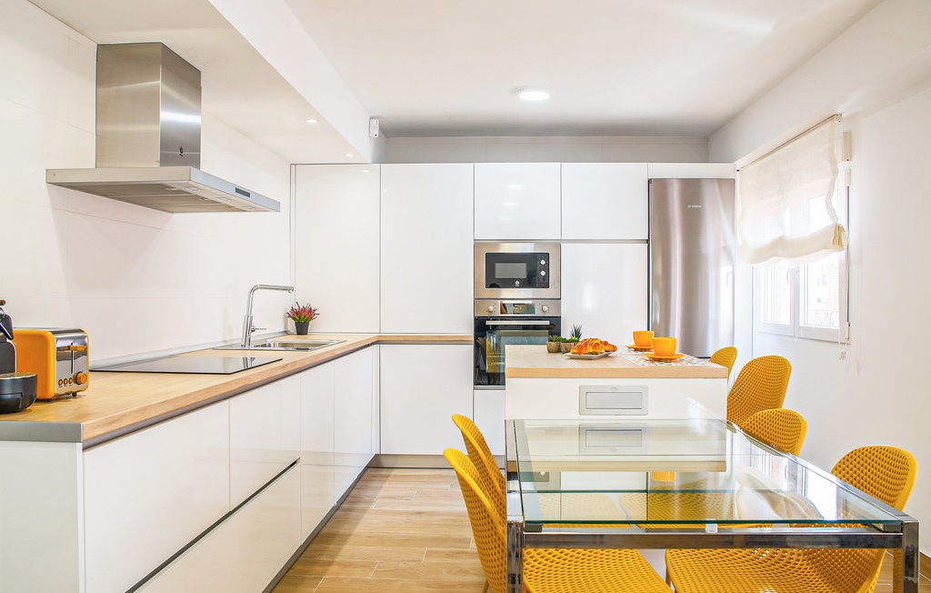 A modern kitchen features sleek white cabinetry and a wooden countertop. A glass dining table with bright yellow chairs is situated in the center, complemented by a well-lit area with natural light flowing through the window.