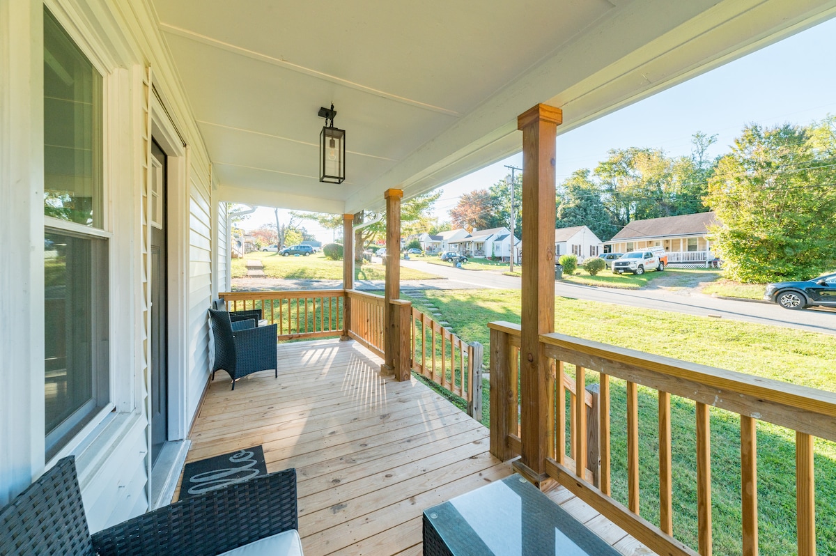 A covered back porch is presented with wooden railings and flooring. Two chairs and a low table are positioned for seating. The view reveals a grassy area and neighborhood street, with several cars parked along the curb.