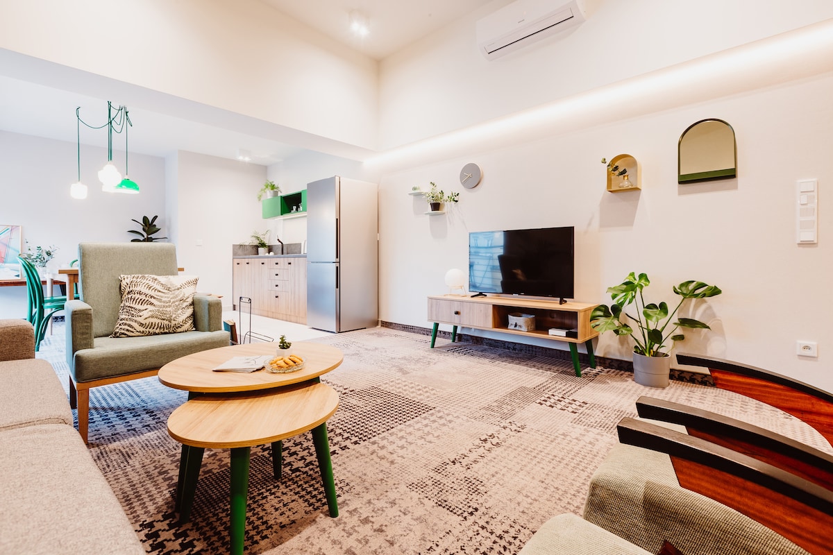 A modern living space features a comfortable seating area with a gray sofa and green accent chairs. A coffee table is set with a snack. An entertainment unit houses a flat-screen TV. The kitchen area is visible, showcasing sleek cabinets and a refrigerator. Natural light illuminates the space.