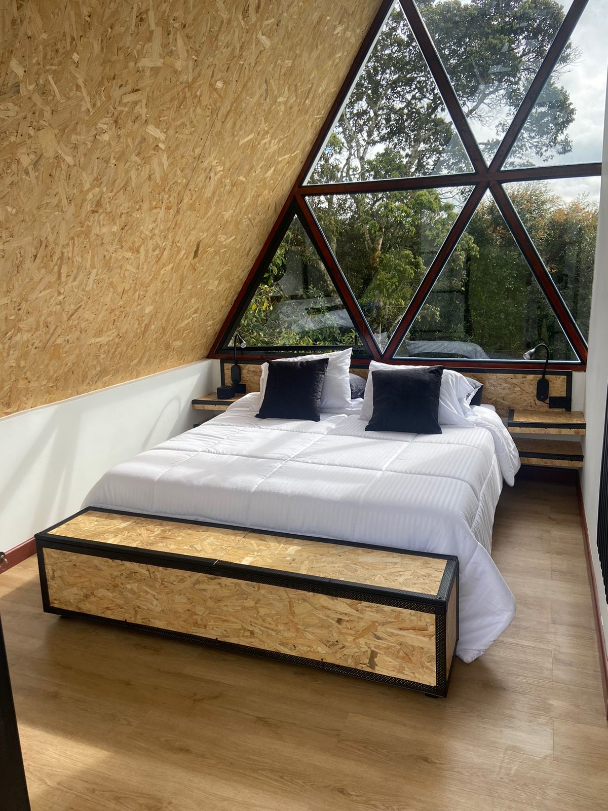 A well-lit bedroom features a spacious bed with white bedding and two black pillows. The walls are made of textured wood, and large triangular windows invite natural light, framing views of the surrounding greenery. Minimalist wooden nightstands are positioned beside the bed.