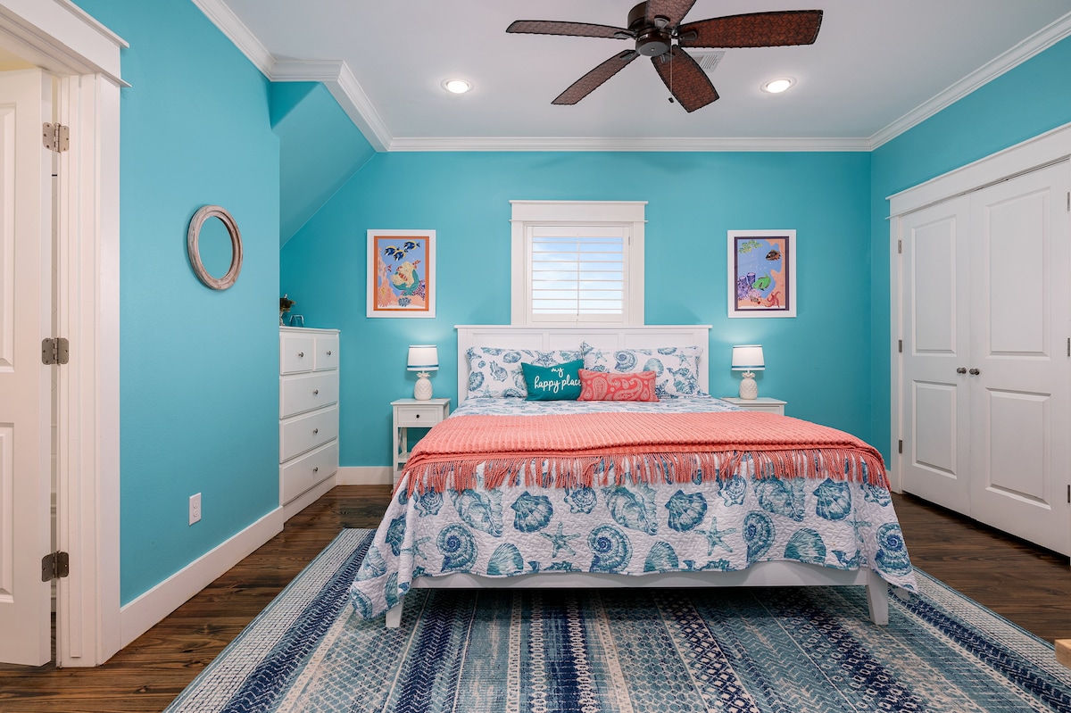 A serene bedroom features bright turquoise walls and a spacious white bed adorned with a coral and blue patterned blanket. Nightstands with lamps flank the bed, while colorful framed art enhances the decor. A large window allows natural light to brighten the room.