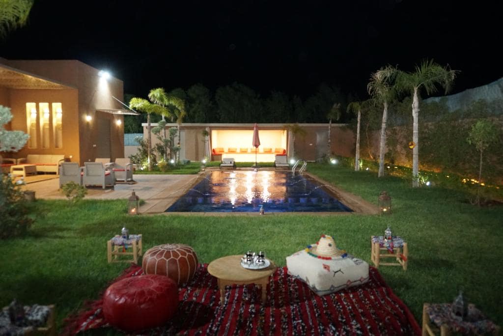 An outdoor area features a private pool surrounded by well-maintained grass. Comfortable seating arrangements are complemented by decorative cushions and small tables. Soft lighting enhances the ambiance, creating a relaxing setting for gatherings at night.