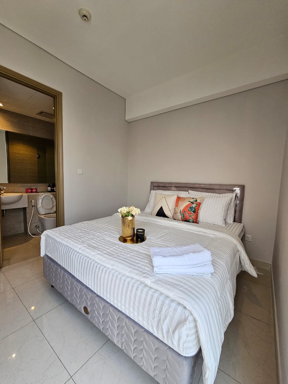 A serene bedroom features a spacious bed adorned with soft linens and decorative pillows. A small table holds a vase of flowers, while light-colored walls and glossy tiles create a bright atmosphere. An ensuite bathroom is conveniently accessible through an open door.