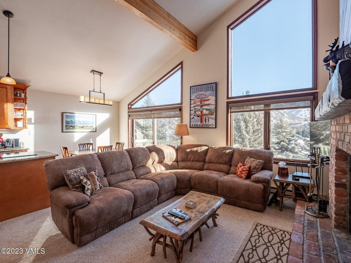 A spacious living area features a large sectional sofa, inviting relaxation. Sunlight filters through multiple windows, highlighting the high ceilings and wooden beams. A rustic coffee table complements the cozy atmosphere, while a brick fireplace adds a touch of warmth to the room.